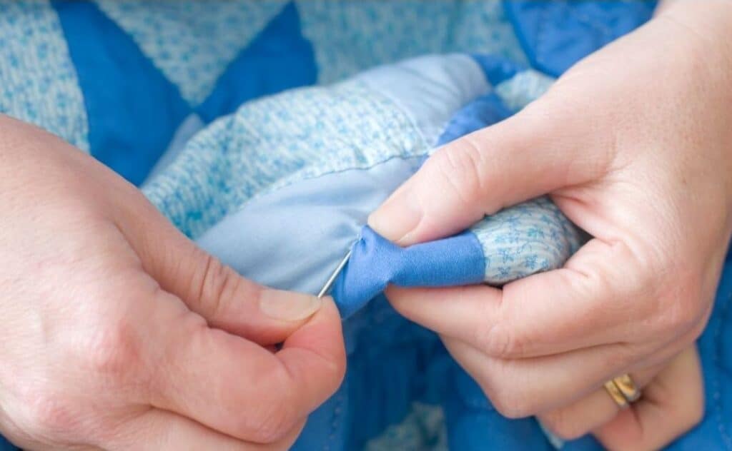 A Mennonites hands carefully stitching a blue patterned quilt, with fine, neat stitches visible. The background shows soft fabric textures.