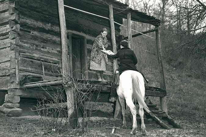 Packhorse librarian on horseback delivering book to woman at log cabin