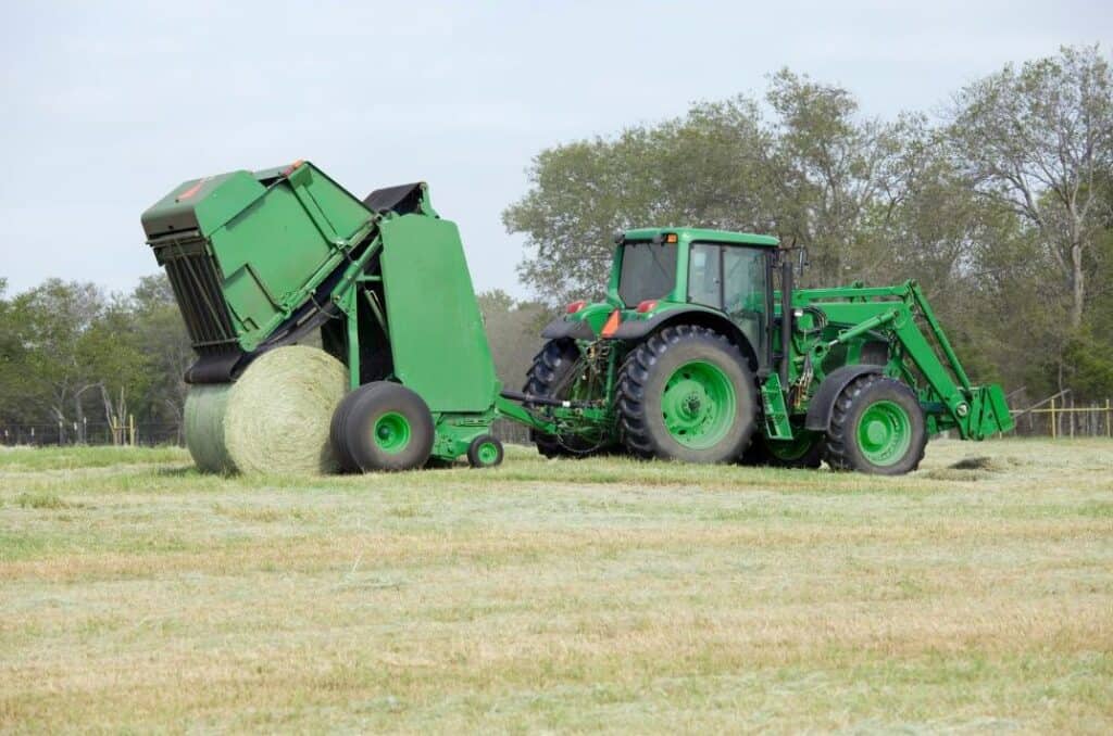 A tractor uses a round baler to lift and release a newly formed hay bale in a cut field.