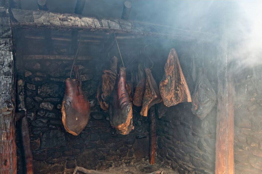 Smoked hams hanging from hooks inside a stone smokehouse.