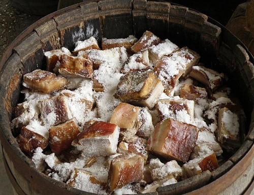Chunks of pork packed in coarse salt inside a wooden curing barrel.