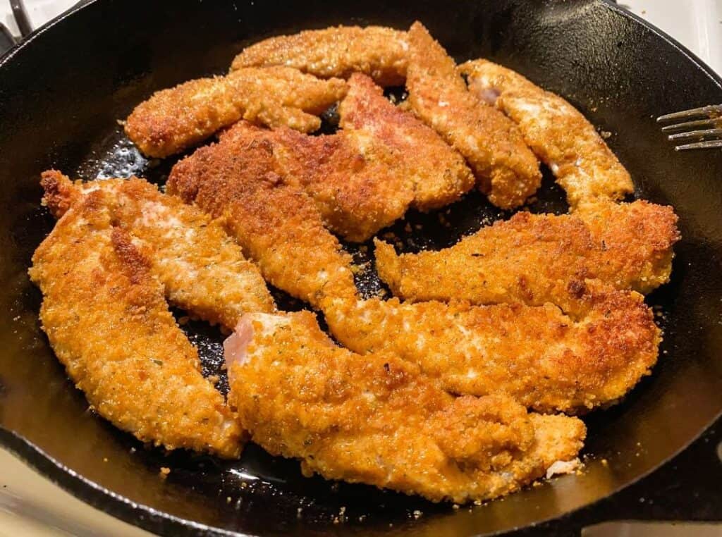 Breaded chicken pieces frying in a cast iron skillet.