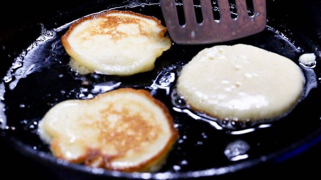 Hoecakes frying in a cast iron skillet with a spatula nearby.