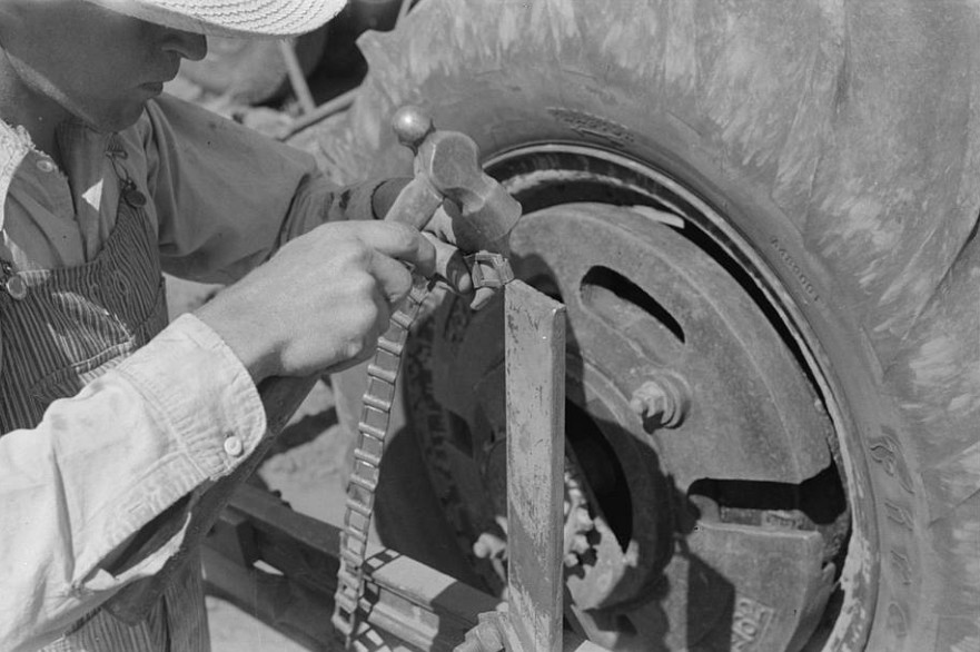 Farm worker repairing a chain link on farm equipment using hand tools. An everyday Appalachian scene often reduced to stereotypes rather than understood in context.