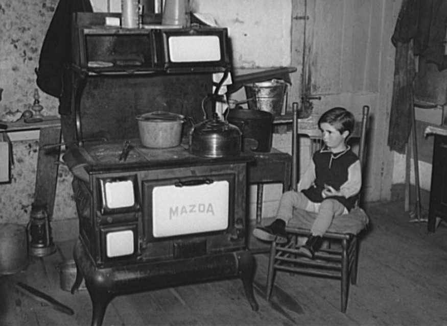 Child seated near a wood stove inside a modest home during winter. An everyday Appalachian scene often reduced to stereotypes rather than understood in context.