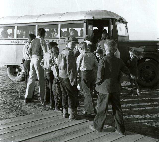 Children boarding a vintage school bus, lined up near the door on a dirt lot.