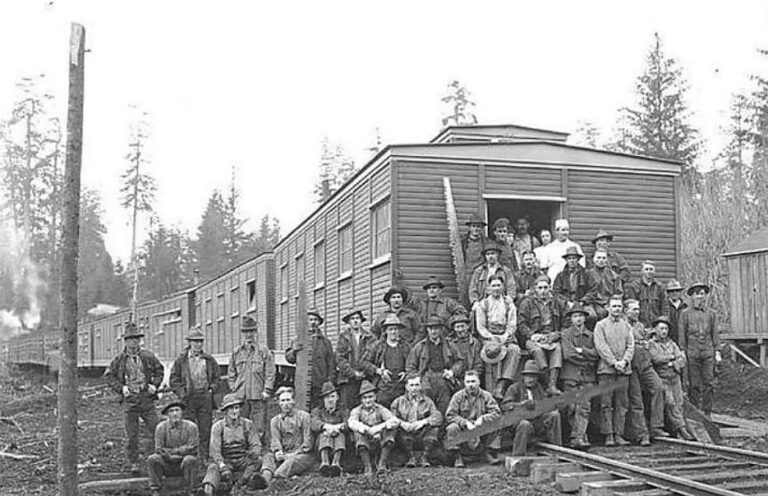 Logging crew gathered beside railroad tracks and camp buildings