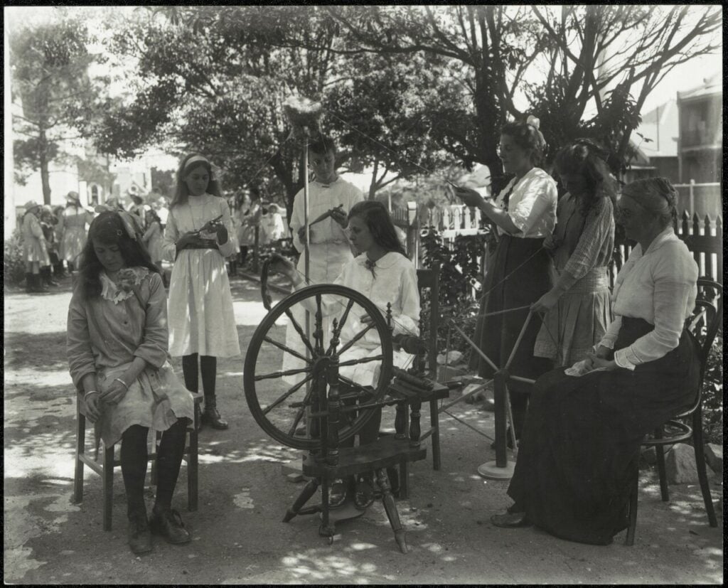 Girls and women gathered around a spinning wheel during a wool-working demonstration
