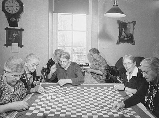 Women seated around a large checkered quilt during a Moravian quilting circle.
