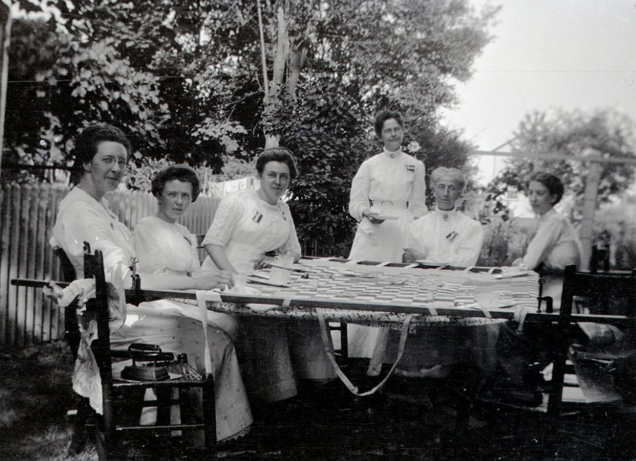 Six women gathered around a full-sized quilt stretched across an outdoor frame.